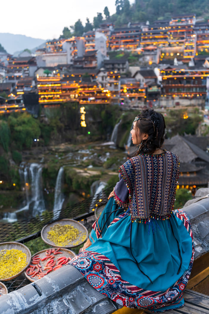 Fotografía de una mujer con vestido tradicional sentada en un tejado, mirando hacia un pueblo antiguo iluminado de noche con una cascada al fondo en China.